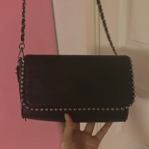 Svart axelväska med nitar och kedja - Cool svart axelväska i skinn med silverfärgade nitar längs kanterna och en axelrem i kedja flätad med läder. Stilren och edgy design som passar perfekt till din outfit när du vill sticka ut lite extra.