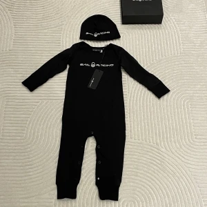 Sail Racing baby set 6–12 mån  - Sail Racing baby set i svart – overall + mössa. Storlek: 6–12 månader. Helt nytt med tagg, aldrig använt. Levereras i originalkartong.