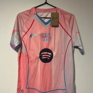 Barça x Nike rosa fotbollströja S - Säljer en FC Barcelona fotbollströja från Nike i rosa och ljusblått med vita linjer och klubbmärke på bröstet. Tröjan har korta ärmar, V-ringning med blå detalj och är tillverkad i lätt Dri-FIT material. Stort svart Spotify-tryck på magen.