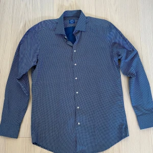 Blå mönstrad skjorta från Whipstitch - Snygg blå skjorta från Whipstitch med små vita mönster över hela tyget. Skjortan har lång ärm, klassisk krage och vita knappar framtill. Skräddarsydd passform som sitter riktigt bra. Perfekt för dig som gillar stilrena detaljer. Storlek 42 och aldrig använd.