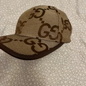 Beige och brun keps från Gucci - Snygg keps från Gucci i beige och brunt med det klassiska GG-mönstret över hela kepsen. Tillverkad i bomull och polyester med justerbar rem bak i brunt. Perfekt accessoar för att lyfta din outfit med en lyxig touch.