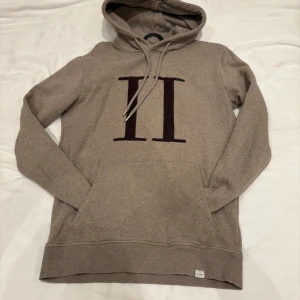 Beige hoodie från Les Deux - Snygg beige hoodie från Les Deux med stor mörkbrun II-logga på bröstet. Tröjan har huva med snörning och en klassisk känguruficka framtill. Tillverkad i mjuk bomullsmix, perfekt för chill dagar eller när du vill ha en clean look.