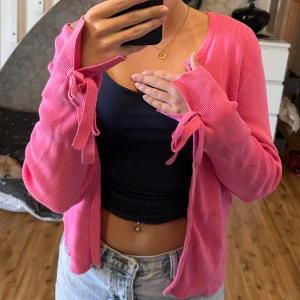 Rosa kofta med knytband - Superfin rosa kofta med långa ärmar och knytband vid ärmsluten. Koftan är i mjuk stickad bomull och har en vågig nederkant som ger en söt touch. Perfekt att slänga över ett linne eller t-shirt för en chill look.