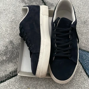 Mörkblå sneakers från Selected Homme - Snygga mörkblå sneakers från Selected Homme i mocka med vit platt sula och svarta snören. Skorna har en stilren design med rund tå och vita detaljer på insidan. Perfekta för dig som gillar en clean och modern look. Använda 2-3 gånger säljer för de känns för stora 