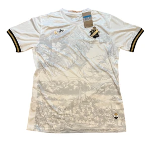 AIK vit matchtröja Nike 2024 - AIK:s officiella vita matchtröja från Nike 2024 med snyggt tryck av Stockholmssilhuett i grått. Tröjan har AIK:s klubbmärke på bröstet, svarta och gula detaljer på ärmsluten och är tillverkad i lätt, ventilerande funktionsmaterial.