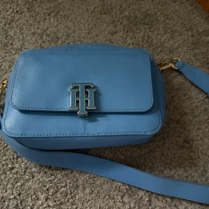 Blå axelväska från Tommy Hilfiger - Snygg ljusblå axelväska från Tommy Hilfiger i skinn med justerbar rem och guldfärgade metalldetaljer. Väskan har ett stort TH-emblem på locket och praktiskt magnetlås. Perfekt storlek för det viktigaste och en stilren accessoar till din outfit.