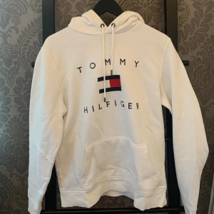 Vit hoodie från Tommy Hilfiger - Snygg vit hoodie från Tommy Hilfiger med klassisk logga och broderad text på bröstet. Tröjan har huva med snörning och en stor magficka. Perfekt för en avslappnad och stilren look. Skriv vid minsta lilla fundering!