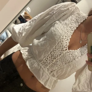 Vit blus med virkade detaljer - Superfin vit blus från F.A.S med virkade spetsdetaljer och pärlknappar längs v-ringningen. Blusen har vida, långa ärmar och resår i midjan för en croppad look. 