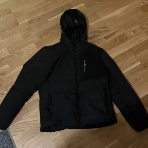 Svart pufferjacka från Sail Racing - Svart pufferjacka från Sail Racing med huva och dragkedja framtill. Jackan har en bröstficka med logga och två sidofickor. Perfekt för kalla dagar och har en stilren look som funkar till det mesta. Materialet är syntet och jackan är vadderad för extra värme. Andvänd i nån månad bara 