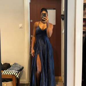 Marinblå balklänning JJ's House - Säljer en marinblå balklänning från JJ's House med smala axelband, v-ringning och hög slits. Klänningen har snygg snörning i ryggen och är tillverkad i ett glansigt, elegant material som ger en lyxig känsla. Perfekt för bal eller festliga tillfällen. Bara använd en gång.