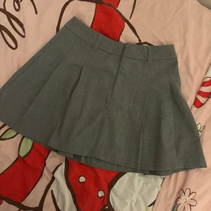 Grå kortkjol från H&M Divided XS - Snygg grå kortkjol från H&M Divided i storlek XS. Kjolen har en klassisk A-linjeform med bred linning och hällor för bälte. Perfekt för dig som gillar en stilren och enkel look. Materialet känns mjukt och har en lätt struktur.