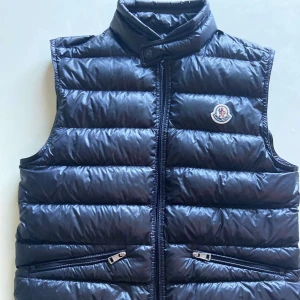Svart dunväst från Moncler - Snygg svart dunväst från Moncler med quiltad design och hög krage. Västen har två fickor med dragkedja framtill och ett broderat Moncler-märke på bröstet. Perfekt att slänga över en hoodie för extra värme och stil. Inga skador. Storlek 1 motsvarar XS-S 100% äkta nypris 8900kr 