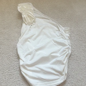 Vit asymmetrisk topp med rosor - Supersnygg vit top med en axel och dekorativa tygrosor på axelbandet. Modellen har rynkade sidor och en elegant, stilren look som verkligen sticker ut!