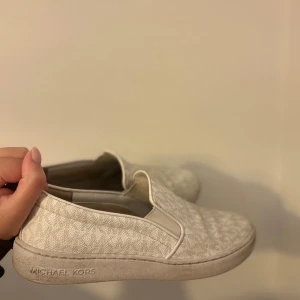 Beige slip-on sneakers Michael Kors - Snygga slip-on sneakers från Michael Kors i beige med diskret mönster och logga på sidan. Skorna har rund tå, platt sula och elastiska paneler för enkel påtagning. Materialet är textil med präglat MK-mönster och små silverdetaljer vid hälen.