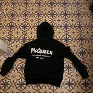 Svart McQueen hoodie med tryck - Svart hoodie från Alexander McQueen med stor vit logotyp och text på bröstet. Tröjan har huva med snörning och långa ärmar. 