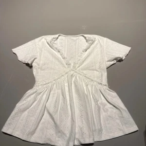 Vit spetsblus från SHEIN - Superfin vit blus från SHEIN med v-ringning och spetsdetaljer längs halsringningen. Blusen har korta ärmar, broderade hålmönster och en lätt utsvängd passform. Perfekt för en fräsch och söt look under varmare dagar.