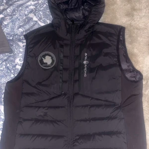 Svart dunväst från Sail Racing - Svart dunväst från Sail Racing med huva och snyggt broderat märke på bröstet. Västen har dragkedja framtill och logotyp på framsidan. Materialet är polyester och dun, vilket gör den både lätt och varm. Bara använt 1 gång då den inte passar. Köpt för 2800kr för 2 år sedan.