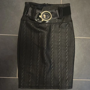 Svart kjol med bälte och guldspänne - Snygg svart kjol med diskret mönster och hög midja. Kjolen har ett brett, glansigt bälte med stort guldspänne som ger en cool statement-look. Materialet är strukturerat och kjolen har en figurnära passform som framhäver siluetten. 