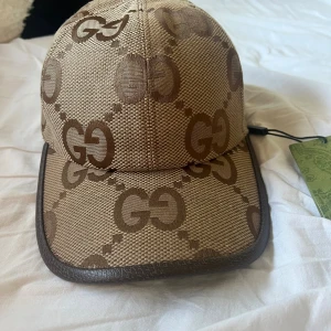 Beige keps från Gucci med GG-mönster - Snygg beige keps från Gucci med klassiskt GG-logomönster i brunt. Kepsen har bruna detaljer i skinn runt skärmen och justerbart band bak. Tillverkad i slitstark canvas och skinn, perfekt för dig som vill sticka ut med en lyxig accessoar.
