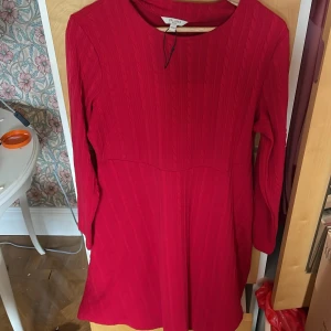 Röd långärmad klänning från Cellbes - Snygg röd klänning från Cellbes i storlek 42/44. Klänningen har lång ärm, rund halsringning och ett subtilt ribbat mönster i tyget. Modellen är kort och har en enkel, klassisk siluett som är lätt att styla.