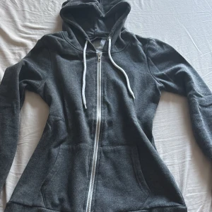 Mörkgrå hoodie med dragkedja - Mysig mörkgrå hoodie med huva och vita snören. Tröjan har hel dragkedja framtill och två stora fickor. Perfekt för chill dagar eller när du vill ha något bekvämt och stilrent på dig.