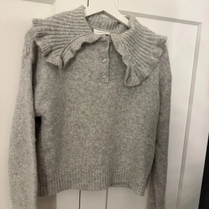 Grå stickad tröja med volangkrage - Superfin grå stickad tröja från Mango med unik volangkrage och knappar framtill. Tröjan har långa ärmar och ribbade muddar vid nederkant och ärmslut. Perfekt för dig som gillar detaljer och vill ha något extra i garderoben.