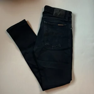 Svarta jeans från Nudie Jeans, 29/30 - Svarta jeans från Nudie Jeans med klassisk femficksdesign och snygga detaljer på bakfickan. Jeansen har en slim passform och är tillverkade i mjukt denimtyg. Perfekta för dig som gillar stilrena och tidlösa byxor.