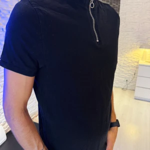 Svart t-shirt med dragkedja - Svart t-shirt med korta ärmar och en cool ståkrage med silverfärgad dragkedja framtill. T-shirten har en clean look och är gjord i mjukt material, perfekt för en stilren och avslappnad vibe. Passar perfekt s/m storlek.