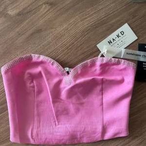 Rosa bandeau tubtopp med strass från NA-KD - Säljer en rosa bandeau tubtopp från NA-KD med glittrande strassdetaljer längs den hjärtformade urringningen. Toppen är croppad och har smockad baksida för extra komfort. Perfekt för dig som vill sticka ut med en färgglad och trendig look.