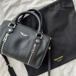 Svart handväska Zadig & Voltaire - Cool svart handväska från Zadig & Voltaire i skinn med silverfärgade detaljer, dubbla dragkedjor framtill och nitar längs kanterna. Väskan har både handtag och axelrem med logga, samt ikonisk vinglogga i metall framtill. Perfekt för dig som gillar edgy och stilren design.