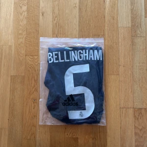 Real Madrid Bellingham tröja Adidas M - Snygg Real Madrid fotbollströja med Bellingham och nummer 5 på ryggen. Tröjan är marinblå med silverfärgade detaljer och Adidas-logga, samt klubbmärke och Emirates Fly Better-tryck på bröstet. Tillverkad i lätt och ventilerande material med korta ärmar.