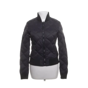 Svart quiltad dunjacka Hollies S - Snygg svart dunjacka från Hollies med quiltat mönster och ribbade muddar vid ärmslut och nederkant. Jackan har dragkedja framtill och står ut med sin bomberjackeform. Perfekt för dig som gillar stilren och sportig look.