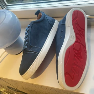 Blå Christian Louboutin sneakers - Snygga blå sneakers från Christian Louboutin i mocka med klassisk röd sula och vita kanter. Skorna har rund tå, platt sula och blå snörning. Insidan är ljusbrun och de har en stilren, minimalistisk design som sticker ut med den ikoniska röda undersidan.