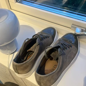 Grå sneakers från Lanvin i mocka - Snygga grå sneakers från Lanvin med ovandel i mocka och glansig tå i mörkgrått. Skorna har vita sulor, rund tå och klassisk snörning. Perfekta för dig som vill ha en stilren och lyxig sneaker med unika detaljer.