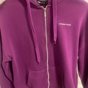 Lila hoodie med dragkedja från new yorker  - Lila hoodie från new yorker med huva, snörning och hel dragkedja framtill. Tröjan har fickor på magen och en liten vit text på bröstet. Perfekt för dig som gillar en chill och färgstark stil.