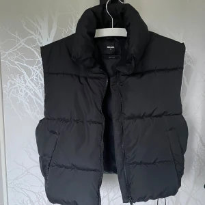 Svart dunväst från Bik Bok - Snygg svart dunväst från Bik Bok med hög krage och dragkedja framtill. Västen har en quiltad design och två stora fickor på sidorna. Perfekt att ha utanpå en hoodie.