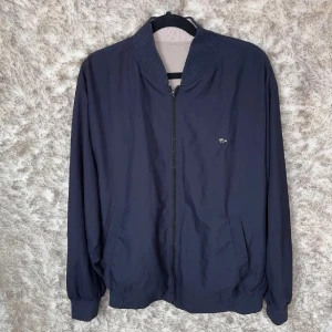 Beige och marinblå bomberjacka Lacoste - Säljer en vändbar bomberjacka från Lacoste i beige och marinblått. Jackan har dragkedja, ribbade muddar och krage samt två fickor på varje sida. Klassisk krokodillogga i metall på bröstet. Perfekt för dig som vill ha två stilar i en jacka.