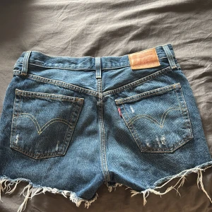 Levi's blå jeansshorts W26 - Säljer då de inte kommer till användning. Blå jeansshorts från Levi's med medelhög midja och fransig kant. Shortsen har slitna detaljer både fram och bak. Perfekta för sommarens alla häng!