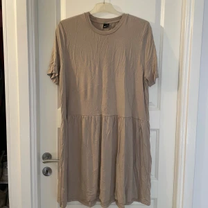 Beige kortklänning från Gina Tricot - En beige kortklänning från Gina Tricot i mjuk bomull. Klänningen har rund hals, korta ärmar och en avslappnad passform med rynkad kjoldel. Perfekt för dig som gillar en enkel och stilren look.