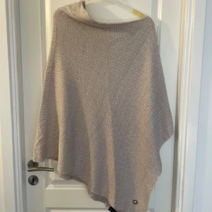 Beige stickad poncho från Stockh LM - Supermysig beige stickad poncho från Stockh LM. Den har en mjuk struktur och är perfekt att slänga över sig när det är kyligt. Ponchon har en lätt asymmetrisk form och är enkel att styla till olika outfits.