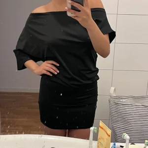 TRENDUG Svart offshoulder fest kort klänning - Snygg svart kort klänning med offshoulder-design och lösare passform upptill. Klänningen har korta ärmar och är figurnära nertill, vilket ger en modern och stilren look. Perfekt för dig som gillar enkel men trendig stil. Helt ny aldrig använd. Köpt på en marknad