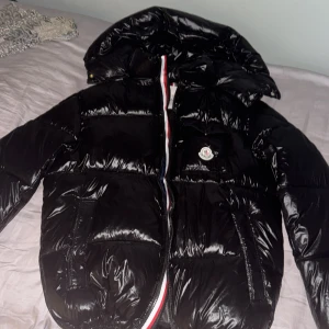 Svart montbelliard pufferjacka från Moncler S - Säljer en svart, glansig pufferjacka från Moncler i storlek S. Jackan har huva, dragkedja med Monclers signaturfärger och logga på bröstet. Perfekt för kalla dagar och riktigt snygg streetstil. Materialet är syntet och dun, vilket gör den både lätt och varm. 