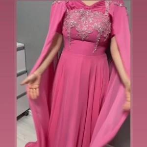 Rosa långklänning med silverbroderi - Säljer en magisk rosa långklänning med svepande capeärmar och vackra silverbroderier över bysten. Klänningen har en elegant, flytande passform och är perfekt för dig som vill sticka ut med en glamorös look. Materialet känns lätt och faller snyggt. Köpte för ett event men slutade med att jag aldrig använt, nu passar den ej längre.  