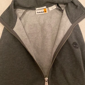 Grå tröja med dragkedja från Timberland - Snygg grå hoodie från Timberland i regular fit med hel dragkedja framtill och broderad logga på bröstet.  Tröjan är tillverkad i mjuk bomullsblandning, perfekt för chill dagar. Klassisk och stilren design som funkar året runt.