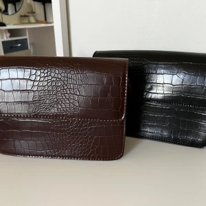 Brunröd och svart axelväska i croco-look - Två stilrena axelväskor i croco-mönstrad syntet, en i mörkbrunt/rött och en i svart. Väskorna har en fyrkantig form, lockstängning och justerbar axelrem. Perfekt för dig som vill ha en trendig och clean accessoar till din outfit. Säljer båda för 200 eller en för 150 🫶🏻