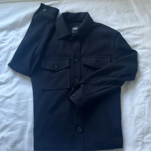 Svart overshirt från Zara - Stilren svart overshirt från Zara med klassisk krage, två stora bröstfickor med knappar och raka ärmar. Jackan har en clean look med knäppning framtill och är perfekt för lager-på-lager. Materialet är mjukt och känns bekvämt mot huden.