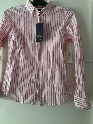 Dam Rand as ostig skjorta från Gina Tricot - Säljer en vit skjorta med rosa ränder från Gina Tricot. Klassisk krage, knäppning framtill och långärmad pryds med diskret broderad logga på bröstet. Tillverkad i tunn bomull som känns lätt och fräsch falling. Perfekt till jeans eller kjol. Kan passa även 36, bröst 94cm.