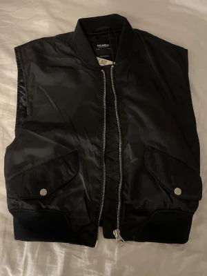 Svart quiltad dunväst Pull&Bear - Säljer en svart quiltad dunväst från Pull&Bear med silkes tyg inuti. Västen har bred mudd nertill som ger en fin passform på kroppen. Två stora fickor med lock och silverfärgade tryckknappar samt en dragkedja framtill. 