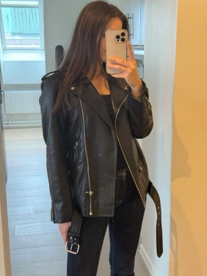 Svart skinnjacka från Stradivarius XS - Säljer en svart skinnjacka från Stradivarius i storlek XS. Jackan har klassisk bikerstil med silvriga dragkedjor, axelklaffar och bälte i midjan. Snygg passform och perfekt att slänga över en hoodie eller t-shirt. Materialet är syntetiskt skinn. Använd 1 gång. 