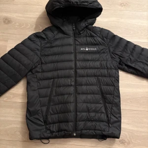 Svart täckjacka från Sail Racing - Säljer en svart tunnare täckjacka från Sail Racing med huva och dragkedja framtill. Jackan har två sidofickor med dragkedja och logga på bröstet samt på huvan. Dragsko längst ner på varje sida. Perfekt för kalla dagar och riktigt snygg till streetwear.